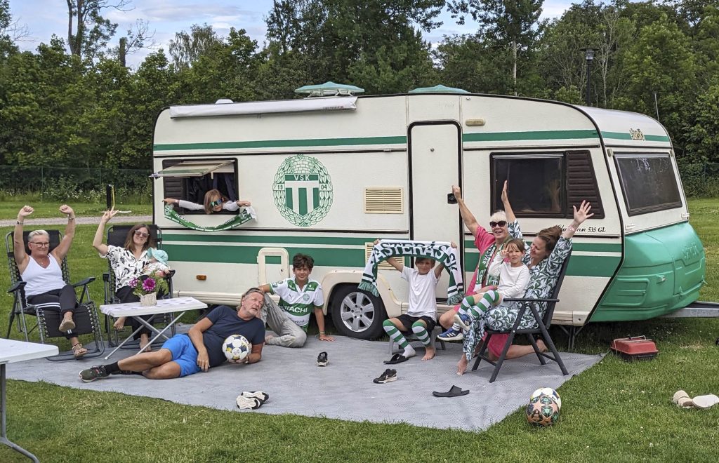 glad familj sitter framför husvagn med grönvita ränder och vsk klubbmärke